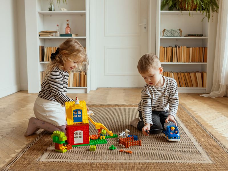 Rodzeństwo buduje razem domek z dużych klocków na dywanie w salonie